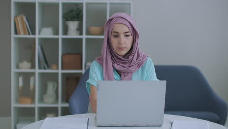 Schöne-Junge-Muslimische-Frau-Arbeitet-Am-Laptop-An-Ihrem-Arbeitsplatz.-Eine-Junge-Muslimische-Frau,-Die-Vor-Einem-Laptop-Bildschirm-Sitzt,-Schaut-Nach-Oben-Und-Blickt-In-Die-Webcam